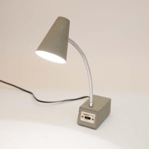 May include: A gray desk lamp with a conical shade and a flexible silver neck attached to a rectangular base. The base features a small black power switch on the front. The lamp is illuminated, casting a bright white light from the shade.