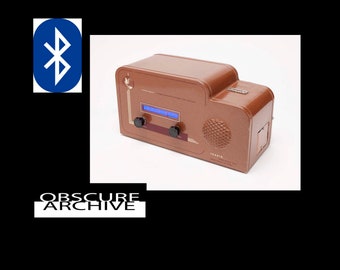 May include: A brown retro-style radio with a blue display screen, two black knobs, and a speaker. The radio has the word "TRADIO" printed on it. A Bluetooth symbol is in the upper left corner. The text "OBSCURE ARCHIVE" is at the bottom.