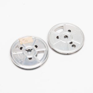 May include: Two silver-colored film reels with a central hub and radial spokes. One reel has a small, rectangular label with handwritten text. The reels are on a white background, suggesting a vintage or retro theme.