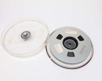 May include: A vintage Scotch audio tape reel with a clear plastic cover. The reel is silver with black tape and a black center hub. The cover is transparent.