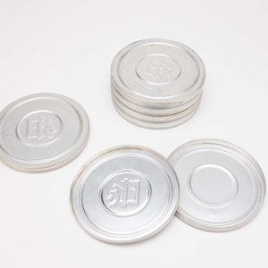 May include: A collection of silver-colored film canisters, some stacked, others scattered. Each lid features embossed text. The canisters are arranged on a white surface, suggesting a vintage or archival theme.