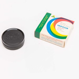 May include: A black film canister and a box of 8mm Indoor Color Film. The film box has a white background with a colorful design of blue, orange, yellow, and red circles. Text on the box reads "8 mm Movie Roll Indoor Color Film 25 Feet."