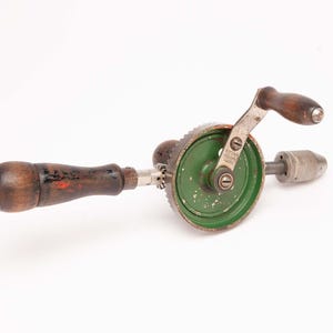 May include: An antique hand drill with a wooden handle and a green gear housing. The drill has a crank handle and a metal chuck for holding drill bits. The wooden handles show signs of wear and age, with some red paint visible.