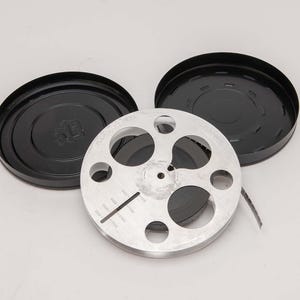 May include: A vintage film reel with a silver metal reel and black film inside, next to two black circular cases. The reel has several circular cutouts and a measurement scale. The cases are open, revealing the reel.