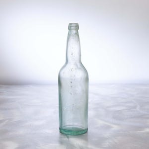 May include: A clear, antique glass bottle with a long neck and rounded body. The bottle has a slight green tint and is empty. The glass shows signs of age, with minor imperfections. The bottle is standing upright.