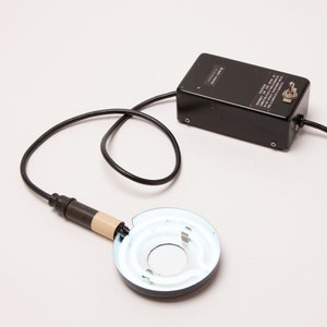 May include: A black device with a circular light at the end of a cord. The device has a black rectangular box with a switch and text. The light is white and circular, with a brown and beige handle.