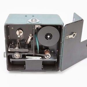 May include: An open, vintage teal-colored film camera with internal mechanisms visible. The camera has a black interior with a film reel, rollers, and a blue tube. The door is open, revealing the inner workings of the device.
