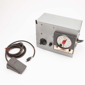 May include: A silver metal timer device with a black dial and a red needle, labeled "TIME-O-LITE." It has a black cord and a gray foot pedal. The device has a knob and a black power cord.