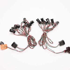 May include: Two sets of vintage Christmas light strings. Each string has multiple black bulb sockets with red and green wires. The strings are bundled with white ties. Each string has a black plug. One plug has an orange tag.