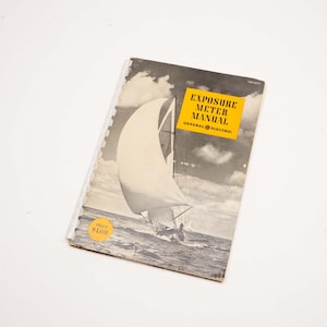 May include: A vintage "Exposure Meter Manual" by General Electric. The cover features a black and white image of a sailboat on the water. A yellow sticker with the text "PRICE $1.00" is on the bottom left corner. The manual is spiral-bound.