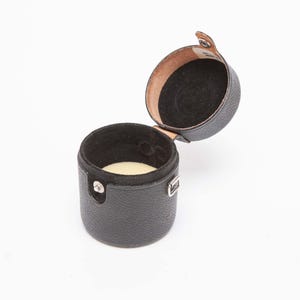 Puede incluir: Un estuche cil&iacute;ndrico negro para reloj con tapa abisagrada. El estuche est&aacute; abierto, revelando un interior negro y un coj&iacute;n color crema. El exterior tiene un acabado texturizado y un cierre plateado.