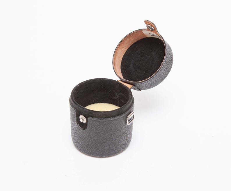 Puede incluir: Un estuche cil&iacute;ndrico negro para reloj con tapa abisagrada. El estuche est&aacute; abierto, revelando un interior negro y un coj&iacute;n color crema. El exterior tiene un acabado texturizado y un cierre plateado.