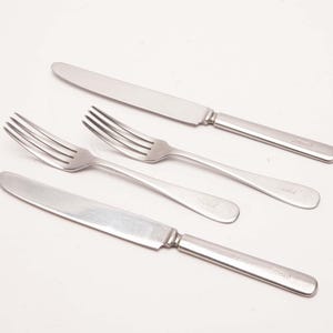 May include: A set of four stainless steel flatware pieces consisting of two dinner knives and two dinner forks. Each piece features a smooth, polished silver finish with a simple, streamlined handle design and a subtle engraved logo detail.