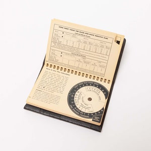 May include: A vintage black and white film exposure calculator with a spiral binding. The open pages display scene index tables and instructions. A circular dial for light and film settings is visible. The calculator is on a white background.