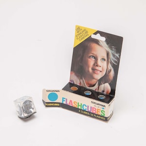 May include: A vintage Sylvania Flashcubes box with a photo of a young girl, and a single flashcube. The box reads "FLASHCUBES" and "3 CUBES/12 FLASHES". The flashcube is a small, clear cube with four bulbs.