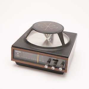 May include: A Zenith solid state radio with a square black base and wood grain trim. The top features a circular black speaker grille with a gold star-like emblem. The front panel displays a horizontal frequency dial and three black control knobs.