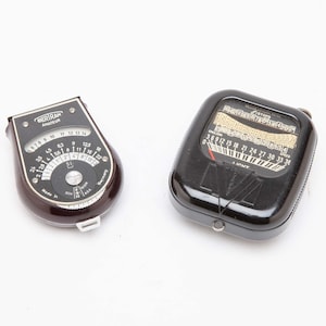 May include: Two vintage light meters on a white background. One is brown and black with the text "BERIRAN AMATEUR". The other is black with a dial and numbers. These devices are used to measure light levels for photography.