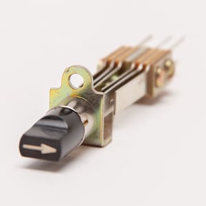 May include: A close-up of a metal toggle switch with a black lever and a white arrow indicating direction. The switch has a gold-colored metal frame and several pins extending from the back. The background is plain white.