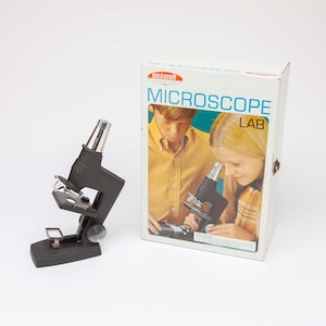 May include: A black microscope with a silver eyepiece and stage is positioned next to a box labeled "MICROSCOPE LAB." The box features an illustration of two children using a microscope. The microscope is a vintage science educational toy.