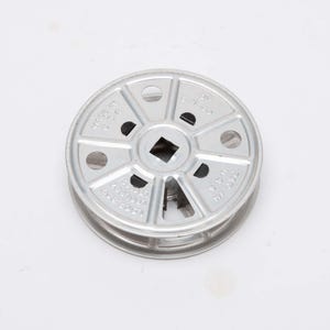 May include: A silver-colored metal reel with a central square opening and circular holes. The reel has text that reads "MADE IN U.S.A." and is likely used for winding or storing something.