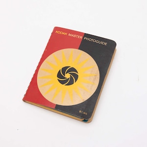 May include: A vintage Kodak Master Photoguide book. The cover is divided into red and black sections, with a yellow and black sunburst design. The book is spiral-bound and has the price $1.95 printed on the bottom right corner.