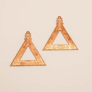 May include: Two bronze-colored, triangular decorative objects with ornate detailing. Each triangle has an open center and a decorative top. The edges are adorned with intricate patterns and symbols, suggesting a historical or symbolic significance.