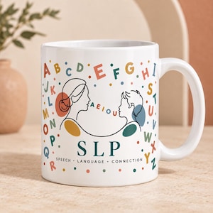 Könnte beinhalten: Ein weißer Keramikbecher mit einer Strichzeichnung eines Erwachsenen und eines Kindes, die sich gegenüberstehen. Das Design enthält das Alphabet in verschiedenen Farben, die Vokale AEIOU und den Text SLP SPEECH LANGUAGE CONNECTION in Dunkeltürkis.