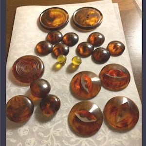 May include: A collection of round buttons in various sizes and shades of amber and brown tortoiseshell patterns. The set includes flat buttons, domed buttons, and two small translucent yellow spherical beads arranged on a white surface.