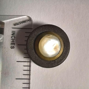 May include: A circular button featuring a dark brown textured outer ring, a gold-toned metal middle band, and a smooth, iridescent white center inlay. A clear plastic ruler marked in inches is positioned along the left side for scale.