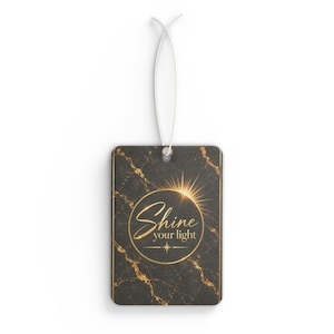 Op de afbeelding: Een rechthoekig hanglabel met een zwart marmerpatroon en gouden aders. Het midden toont een goudkleurig cirkelvormig embleem met een zonnestraalgrafiek en de tekst "Shine your light" in cursief schrift, opgehangen aan een wit koord.