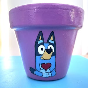 May include: A lilac-coloured flower pot decorated with a cartoon dog holding a red heart. The dog is blue, black, and white. The pot is a standard terracotta shape, ideal for planting flowers or herbs.
