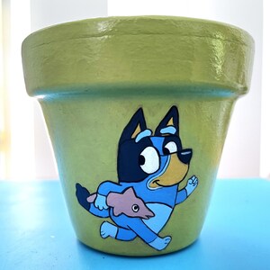 May include: A hand-painted, pale green ceramic plant pot with a cartoon dog character in blue, black, and yellow. The dog is running and holding a blue fish. The pot is on a blue surface.
