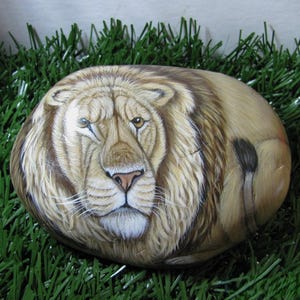 May include: A hand-painted rock featuring a lion with a light brown mane and a light brown body. The lion is painted with realistic details, including its fur, eyes, and whiskers. The rock is resting on a bed of green grass.