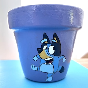 May include: A hand-painted, lavender ceramic plant pot with a cartoon character in blue, black, and tan. The character is in a playful pose, with one leg raised. The pot is suitable for small plants and measures approximately 10 cm in diameter.