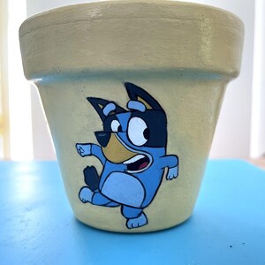 May include: A hand-painted, pale yellow ceramic plant pot with a cartoon character in blue, black, and white. The character is running with an open mouth. Suitable for indoor or outdoor use, this pot is ideal for plants.