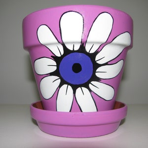 May include: A pink painted flower pot with a matching saucer. The pot features a large white daisy design with a blue centre and black outline. The pot is designed for indoor or outdoor use.