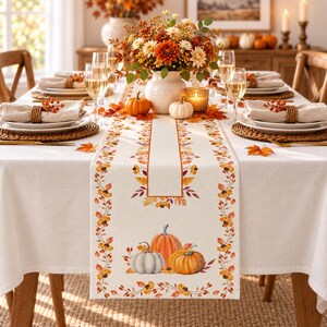 May include: A cream-colored fabric table runner featuring a printed border of autumn leaves, acorns, and berries, with a central illustration of three pumpkins in orange and white, designed for a dining table setting with seasonal decor.