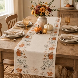 May include: A cream-coloured table runner featuring a printed pattern of orange pumpkins, brown acorns, and various autumn leaves. The runner is displayed on a wooden dining table set with white plates, beige napkins, gold cutlery, and amber glassware.