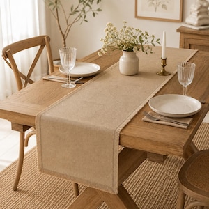 Puede incluir: Un camino de mesa rectangular de color beige con borde deshilachado sobre una mesa de comedor de madera. El camino está centrado en la mesa, que está puesta con dos platos blancos, dos vasos transparentes estriados, un florero blanco y una vela.