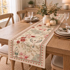 Può includere: Un runner da tavola color crema con una stampa botanica di fiori rossi, foglie verdi e uccelli. Il runner presenta un bordo decorativo con un motivo floreale ripetuto ed è disposto su un tavolo da pranzo in legno con stoviglie.