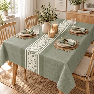 May include: A sage green textured tablecloth with a central white botanical leaf print runner, set with round woven placemats, white ceramic plates, linen napkins with wooden rings, clear glassware, and two pillar candles in woven holders.