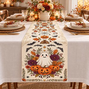 May include: A cream-coloured table runner featuring a repeating pattern of orange pumpkins, black bats, spider webs, moths, and a central illustration of a white ghost surrounded by orange and purple flowers and three jack-o'-lanterns.