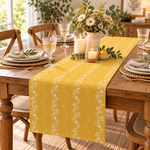 Puede incluir: Un camino de mesa amarillo mostaza con un patrón repetitivo de enredaderas florales blancas y líneas onduladas verticales. El camino está colocado sobre una mesa de comedor de madera con platos, servilletas, cristalería, un florero y dos velas.