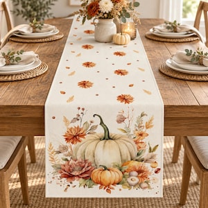 Puede incluir: Un camino de mesa de color crema con un estampado de acuarela que muestra una calabaza blanca grande, una calabaza naranja más pequeña y flores de otoño, incluyendo dalias y follaje seco, con pequeños motivos florales naranjas.