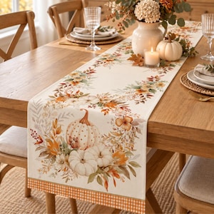 Puede incluir: Un camino de mesa rectangular con un patrón estampado de calabazas blancas, hojas de otoño naranjas y marrones, y elementos florales. El camino tiene un fondo color crema y un borde con un pequeño estampado de cuadros naranjas y blancos.