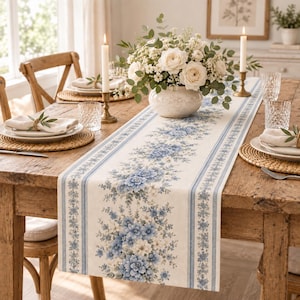 Puede incluir: Un camino de mesa rectangular largo de tela color crema con un patrón central de racimos de flores azules y blancas, bordeado por finas rayas azules. El camino de mesa está colocado sobre una mesa de madera con vajilla y un florero.