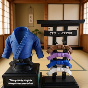 Pode incluir: Um top de gi de jiu-jitsu azul sobre uma base preta com o texto "Todo grande projeto começa com uma Ideia" e um suporte preto estilo torii segurando faixas marrom, roxa, azul e branca com o texto "JIU - JITSU" na barra transversal.