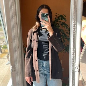 May include: A dark brown suede jacket featuring a classic collar, long sleeves, and a button-down front closure. The jacket is worn over a black mock neck top and light blue denim jeans, creating a layered look with visible stitching details.