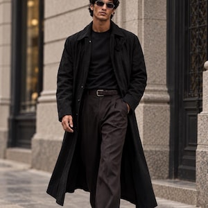 May include: A long black trench coat worn over a black t-shirt and dark brown trousers with a belt. The outfit includes dark-tinted sunglasses and black leather loafers. The ensemble features a structured collar, long sleeves, and a straight-leg cut.