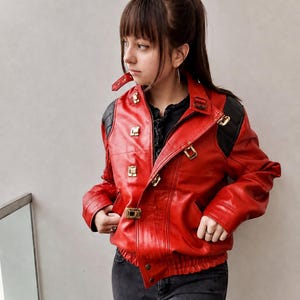 Peut inclure: Une veste en cuir rouge vif avec des empiècements d'épaules matelassés noirs, des boucles métalliques dorées et une fermeture éclair frontale. La veste présente un col haut avec une bride à boucle, des manches longues et un ourlet élastiqué.